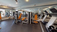 This fitness center is a comprehensive, single-floor facility designed to support a full range of workout routines. The space is outfitted with an extensive selection of strength-training machines accented in distinctive orange and silver, including cable machines, leg press, chest press, and a variety of selectorized equipment. A well-organized dumbbell rack provides a full range of free weights, catering to users of all fitness levels. Cardio equipment, including treadmills and elliptical machines, lines one section of the floor, offering options for aerobic exercise alongside the strength machines. Floor-to-ceiling mirrors span multiple walls, visually expanding the space and allowing users to monitor their form throughout their workouts. The dark rubber flooring provides a durable, functional surface suited for high-traffic gym use. Overhead, multiple ceiling fans complement the recessed lighting, ensuring comfort and consistent airflow throughout the room. Large windows along one wall invite natural light and offer views of the surrounding exterior. French doors provide easy access points on either end of the space. A wall-mounted flat-screen television adds an entertainment element for those seeking a more engaging workout experience. The overall atmosphere is clean, organized, and purpose-built for an effective fitness experience.