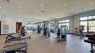 This single-floor fitness and wellness facility is thoughtfully designed to support a comprehensive health and exercise experience. The main workout area is generously proportioned, featuring soft, durable flooring throughout and bathed in natural light from large multi-pane windows that offer views of the surrounding greenery. Recessed ceiling lighting supplements the natural light, creating a bright and energizing atmosphere.

The gym floor is outfitted with an extensive array of Matrix-brand cardio equipment, including treadmills, elliptical trainers, and stationary bikes, arranged in organized rows. A full selection of strength-training machines, cable systems, and free weights lines the walls and center floor. Floor-to-ceiling mirrors along one wall visually expand the space and assist with form. A wall-mounted flat-screen TV and a built-in water fountain add practical convenience.

French doors with wall sconces provide a welcoming entrance, connecting to a foyer with elegant pendant lighting. A dedicated treatment room offers a serene spa-like retreat, featuring a massage table with plush linens, white subway tile wainscoting, soft sage-green upper walls, and calming artwork. An attached vanity area includes a vessel sink set against marble-style tile, built-in white cabinetry, and open shelving stocked with amenities, completing the wellness-focused environment.