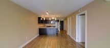 This single-floor space offers a thoughtfully designed open-concept layout that seamlessly blends the kitchen and living areas into one cohesive environment. The warm-toned hardwood flooring runs throughout, lending a sense of continuity and warmth to the space.

The kitchen is a standout feature, showcasing rich dark espresso cabinetry that contrasts elegantly with the light stone countertops. A peninsula-style island provides additional workspace and casual seating, making it ideal for both cooking and entertaining. Stainless steel appliances, including a refrigerator and microwave, add a modern, polished touch. Three stylish pendant lights hang above the workspace, providing focused task lighting while contributing to the overall contemporary aesthetic. Track lighting further complements the kitchen's illumination.

The open living area benefits from the natural flow of the layout, creating a spacious and inviting atmosphere. Neutral warm-toned walls throughout enhance the sense of openness and pair harmoniously with the flooring. A hallway leads to additional rooms, with white panel doors providing clean, classic transitions between spaces. The overall ambiance is modern yet welcoming, combining functional design with contemporary finishes for a comfortable and well-appointed living environment.