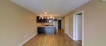 This single-floor space offers a thoughtfully designed open-concept layout that seamlessly blends the kitchen and living areas into one cohesive environment. The warm-toned hardwood flooring runs throughout, lending a sense of continuity and warmth to the space.

The kitchen is a standout feature, showcasing rich dark espresso cabinetry that contrasts elegantly with the light stone countertops. A peninsula-style island provides additional workspace and casual seating, making it ideal for both cooking and entertaining. Stainless steel appliances, including a refrigerator and microwave, add a modern, polished touch. Three stylish pendant lights hang above the workspace, providing focused task lighting while contributing to the overall contemporary aesthetic. Track lighting further complements the kitchen's illumination.

The open living area benefits from the natural flow of the layout, creating a spacious and inviting atmosphere. Neutral warm-toned walls throughout enhance the sense of openness and pair harmoniously with the flooring. A hallway leads to additional rooms, with white panel doors providing clean, classic transitions between spaces. The overall ambiance is modern yet welcoming, combining functional design with contemporary finishes for a comfortable and well-appointed living environment.