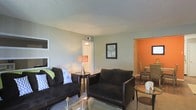 This single-floor residence offers a thoughtfully arranged layout visible from the living room, which serves as the central gathering space. The living room is furnished with dark leather-upholstered sofas accented with decorative throw pillows, arranged around a glass-topped coffee table that adds a light, open feel to the area. Soft, neutral green-gray walls create a calm backdrop, while a textured ceiling adds subtle architectural character. A framed wall print and a warm-toned table lamp contribute to the room's inviting ambiance. The flooring appears to be a neutral carpet, reinforcing the comfortable atmosphere. An HVAC vent is integrated into the ceiling, ensuring climate comfort throughout the space. Adjacent to the living room, an open passageway leads to a dining area distinguished by a vibrant orange accent wall, creating a striking visual contrast and adding energy to the space. Dining chairs are partially visible, suggesting a dedicated eating area. A doorway from the living room leads further into the residence, hinting at additional rooms beyond. Overall, the space balances a relaxed, functional aesthetic with thoughtful use of color and furnishings.