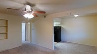 This single-floor residence offers a straightforward and functional layout designed for comfortable everyday living. The living area is finished with wall-to-wall carpeting that adds warmth underfoot, while a ceiling fan with an integrated light fixture provides both air circulation and ambient lighting overhead. Neutral-toned walls throughout create a clean, versatile backdrop suitable for a variety of décor styles.

The living space flows openly into the kitchen, which is accessible through a wide arched passageway. The kitchen features white upper cabinetry that brightens the space and a full-size black refrigerator that serves as a bold contrast against the lighter surroundings. The kitchen floor transitions to a hard surface, distinguishing it from the carpeted living zone.

A hallway visible from the main living area leads to additional rooms, suggesting a practical and well-connected floor plan. The presence of a thermostat on the living room wall indicates individual climate control for the unit. Recessed vents are positioned in the ceiling for efficient air distribution throughout. Overall, the space presents a clean and livable environment with a no-frills, functional aesthetic that makes efficient use of its single-level footprint.