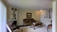 This single-floor residence offers a comfortable and classically appointed living environment. The living room serves as the heart of the space, showcasing rich dark hardwood flooring that transitions to a plush area rug beneath the seating arrangement. Crown molding frames the textured ceiling, adding a refined architectural detail that enhances the room's character. Warm, neutral-toned walls provide a welcoming backdrop, complemented by carefully selected furnishings including a tufted sofa and an ornate wooden coffee table with wrought iron accents. A framed landscape painting serves as a tasteful focal point on the main wall, while decorative wall art and sculptural pieces add depth and personality to the space. A fireplace is visible along one wall, contributing to the room's cozy ambiance. Natural and ambient lighting work together to create a warm, inviting atmosphere throughout. An entry door and adjacent areas are visible, hinting at a well-connected floor plan. Overall, this residence combines classic design elements with comfortable, functional living spaces to create a cohesive and welcoming home environment.