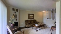 This single-floor residence offers a comfortable and classically appointed living environment. The living room serves as the heart of the space, showcasing rich dark hardwood flooring that transitions to a plush area rug beneath the seating arrangement. Crown molding frames the textured ceiling, adding a refined architectural detail that enhances the room's character. Warm, neutral-toned walls provide a welcoming backdrop, complemented by carefully selected furnishings including a tufted sofa and an ornate wooden coffee table with wrought iron accents. A framed landscape painting serves as a tasteful focal point on the main wall, while decorative wall art and sculptural pieces add depth and personality to the space. A fireplace is visible along one wall, contributing to the room's cozy ambiance. Natural and ambient lighting work together to create a warm, inviting atmosphere throughout. An entry door and adjacent areas are visible, hinting at a well-connected floor plan. Overall, this residence combines classic design elements with comfortable, functional living spaces to create a cohesive and welcoming home environment.