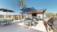 Located at 6900 S 59th Ave in Phoenix, Arizona, this single-level outdoor amenity space is thoughtfully designed to maximize the comfort and lifestyle of its residents in the Sonoran Desert climate. The centerpiece of the space is a resort-style swimming pool, enclosed by sleek metal fencing and framed by towering palm trees that lend a tropical ambiance to the surroundings.

A generously sized covered pergola area provides a shaded outdoor lounge, furnished with plush navy blue sectional sofas, coordinating accent pillows, and contemporary white coffee tables — creating an inviting space for relaxation and socializing. Adjacent to the lounge, an open-air dining patio is outfitted with multiple tables and chairs shaded by large blue umbrellas, ideal for outdoor meals and gatherings under the Arizona sun.

A secondary covered patio area, equipped with a ceiling fan and recessed lighting, offers a more intimate dining setting with direct views of the pool. A private balcony-style lounge features a sculptural stone coffee table and an L-shaped sectional sofa, offering a quieter retreat with views of the community.

Throughout the space, light-toned tile and concrete flooring provide a clean, modern aesthetic that complements the neutral stucco architectural elements. The overall ambiance is polished yet relaxed, blending resort-style comfort with functional, community-oriented outdoor living.