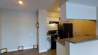 This single-floor residence offers a practical and well-organized layout designed for comfortable everyday living. The kitchen is a standout feature, equipped with black appliances including a full-size refrigerator and an electric range with overhead ventilation. White cabinetry lines the upper walls, providing ample storage space, while the countertops feature a granite-style surface that adds a touch of refinement to the functional design. A breakfast bar or partial peninsula extends from the kitchen, offering additional prep space and a natural transition into the adjacent living area. The open sightlines between the kitchen and living space create a sense of connectivity throughout the floor plan, making the space feel larger and more inviting. Warm-toned wood-look flooring runs throughout, adding warmth and visual continuity. The walls are finished in a neutral light gray, contributing to a clean and modern aesthetic. Practical details such as a mounted fire extinguisher reflect attention to safety. Overall, this single-floor space combines functionality with a straightforward, modern design, making it well-suited for those seeking an efficient and cohesive living environment.