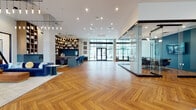 This elegantly designed single-floor amenity space offers a seamless blend of modern architecture and curated interior design. The expansive open floor plan is anchored by striking herringbone-patterned hardwood flooring that flows consistently throughout every zone of the space. Soaring ceilings with cascading pendant light fixtures create a dramatic vertical presence, enhancing the sense of openness and grandeur.

A standout feature is the glass-enclosed conference room, framed in sleek dark metal, which provides a private meeting area while maintaining visual connectivity to the surrounding lounge. Adjacent to this, a comfortable seating area is anchored by a linear gas fireplace set against a dark steel surround, with built-in wood shelving above and plush armchairs arranged for conversation.

The coffee and refreshment bar showcases a geometric blue tile backsplash, warm wood cabinetry, and marble-look countertops. Floor-to-ceiling windows and glass entry doors flood the interior with natural light and offer direct views to an outdoor pool and courtyard area. A dedicated lounge nook features leather sofas, an organic wood coffee table, and bold wall art beneath dramatic pendant lighting. The overall ambiance is refined, contemporary, and highly functional.