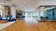 This elegantly designed single-floor amenity space offers a seamless blend of modern architecture and curated interior design. The expansive open floor plan is anchored by striking herringbone-patterned hardwood flooring that flows consistently throughout every zone of the space. Soaring ceilings with cascading pendant light fixtures create a dramatic vertical presence, enhancing the sense of openness and grandeur.

A standout feature is the glass-enclosed conference room, framed in sleek dark metal, which provides a private meeting area while maintaining visual connectivity to the surrounding lounge. Adjacent to this, a comfortable seating area is anchored by a linear gas fireplace set against a dark steel surround, with built-in wood shelving above and plush armchairs arranged for conversation.

The coffee and refreshment bar showcases a geometric blue tile backsplash, warm wood cabinetry, and marble-look countertops. Floor-to-ceiling windows and glass entry doors flood the interior with natural light and offer direct views to an outdoor pool and courtyard area. A dedicated lounge nook features leather sofas, an organic wood coffee table, and bold wall art beneath dramatic pendant lighting. The overall ambiance is refined, contemporary, and highly functional.