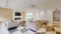 Located at 507 E Main St in Bound Brook, NJ, this single-floor space showcases a refined, contemporary design with high-end finishes throughout. The main lounge area is a standout, featuring a striking geometric marble-tile floor in contrasting light and dark tones, a sleek linear electric fireplace set against a warm wood-panel accent wall, and a wall-mounted flat-screen television. Plush neutral-toned seating arrangements create an inviting, comfortable atmosphere, while a round glass coffee table adds a touch of sophistication. A dedicated dining nook is anchored by a round glass-top table with gold-accented legs, surrounded by elegant upholstered chairs and illuminated by a striking circular crystal chandelier. Natural light fills the space through multiple large windows dressed with clean roller shades, complemented by gold wall sconces that add a warm, ambient glow. The kitchenette area is outfitted with crisp white shaker cabinetry, white quartz countertops, a subway tile backsplash, a stainless steel sink, and an under-counter beverage refrigerator. Additional cabinetry and prep space provide generous storage and utility. The overall ambiance is bright, airy, and polished, making this a versatile and visually impressive space.