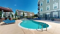 Located at 7500 Hearthside Way in Elkridge, Maryland, this outdoor amenity space serves as the central gathering hub of a multi-story residential community. The centerpiece is a generously sized, freeform swimming pool with smooth curved edges and a dedicated entry staircase with stainless steel handrails, complemented by an adjacent spa for relaxation. Surrounding the pool is an expansive concrete sun deck furnished with rows of blue lounge chairs and shaded seating areas beneath navy blue market umbrellas, providing ample space for leisure and socializing. A separate courtyard level, accessible via stone-flanked steps with ornamental iron railings, features an outdoor grilling station and additional patio seating, making it well-suited for outdoor entertaining. The entire area is enclosed by a decorative black iron security fence and set against a backdrop of the community's multi-story buildings, which display a classic architectural style combining brick and siding exteriors with private balconies overlooking the pool area. Mature trees and landscaped greenery add natural contrast throughout the space. The two-level layout of this amenity area offers distinct zones for active recreation and relaxed gathering, all bathed in open sky and natural sunlight.