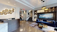 This elegantly designed lobby serves as a refined entryway, combining contemporary aesthetics with high-end materials to create a welcoming, upscale atmosphere. The gleaming terrazzo floors extend throughout the entire space, reflecting light and lending a polished, luxurious feel underfoot.

At the entrance, a dramatic marble-clad vestibule with brass wall sconces and large windows frames the glass entry doors, allowing natural light to pour into the space. The main lobby features a curved, dark-toned reception desk with a marble front panel, positioned beneath a sculptural brass wall installation that adds an artistic focal point. Adjacent to the desk, a bank of individual mailboxes is neatly integrated into a dark built-in unit.

A comfortable lounge area anchors one side of the lobby, featuring a rich navy velvet sofa, a coordinating accent chair, and gold-toned glass coffee tables arranged atop a soft area rug. A dark built-in shelving unit displays curated décor alongside a framed abstract artwork. Pendant lighting with gold interiors adds warmth throughout. A staircase with sleek dark treads and a black handrail, along with an elevator, provides access to upper levels, completing this thoughtfully appointed lobby.