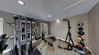 This single-floor fitness space is designed to accommodate a range of workout needs within a compact, functional layout. The room features neutral-toned walls in shades of gray and beige, complemented by clean white baseboards and a smooth white ceiling with recessed lighting that ensures the space is well-illuminated throughout. The flooring consists of a low-pile, textured carpet in a warm beige tone, providing a degree of cushioning underfoot. A wall-mounted mirror on one side of the room enhances the sense of space and allows users to monitor their form during exercise. The equipment selection is diverse, including a multi-station cable and weight machine along the left wall, an elliptical trainer, a rowing or ab machine on the floor, a water-filled freestanding cooler, and a vertical knee raise and dip station positioned toward the right side of the room. The overall ambiance is clean and utilitarian, reflecting a straightforward approach to fitness facility design. The space is practical and organized, making it suitable for a variety of strength and cardio-focused training activities.