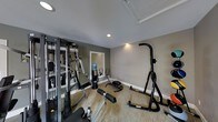 This single-floor fitness space is designed to accommodate a range of workout needs within a compact, functional layout. The room features neutral-toned walls in shades of gray and beige, complemented by clean white baseboards and a smooth white ceiling with recessed lighting that ensures the space is well-illuminated throughout. The flooring consists of a low-pile, textured carpet in a warm beige tone, providing a degree of cushioning underfoot. A wall-mounted mirror on one side of the room enhances the sense of space and allows users to monitor their form during exercise. The equipment selection is diverse, including a multi-station cable and weight machine along the left wall, an elliptical trainer, a rowing or ab machine on the floor, a water-filled freestanding cooler, and a vertical knee raise and dip station positioned toward the right side of the room. The overall ambiance is clean and utilitarian, reflecting a straightforward approach to fitness facility design. The space is practical and organized, making it suitable for a variety of strength and cardio-focused training activities.
