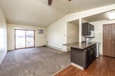 This single-floor unit offers a practical and well-organized layout across a comfortable footprint. The living area is a notable highlight, featuring vaulted ceilings with a textured finish that amplify the sense of space. A ceiling fan provides air circulation, while a sliding glass door opens directly onto a private balcony, filling the room with natural light and offering outdoor views. Baseboard heating runs along the perimeter, ensuring consistent warmth throughout. A peninsula with granite-style countertops and dark cabinetry separates the living space from the kitchen, which is laid out in a galley configuration. The kitchen features dark lower and upper cabinets, laminate stone countertops, white appliances including a range, microwave, and dishwasher, and warm wood-tone flooring. A pantry closet provides additional storage. The bedrooms are carpeted and each equipped with a ceiling fan and a window that brings in natural light, along with baseboard heating. Two bathrooms serve the unit: one features a vanity with stone-style countertops and a tub-shower combination with sliding glass doors and wood-tone flooring, while the second offers a vanity with matching finishes and a sliding closet door nearby.