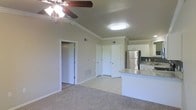 This single-floor unit features an open-concept layout that seamlessly connects the living area to the kitchen. The living space is finished with plush carpet flooring and accented by crown molding along the upper walls, adding a polished touch to the overall aesthetic. A ceiling fan with a built-in light fixture is centrally positioned, providing both comfort and illumination to the space.

The kitchen is situated adjacent to the living area and is separated by a peninsula counter finished with granite-style laminate countertops, offering convenient casual seating or prep space. White cabinetry lines the kitchen walls, complemented by a stainless steel refrigerator and a tile backsplash, creating a clean, modern look. The kitchen flooring transitions to light-toned ceramic tile, providing a durable, easy-to-clean surface.

Multiple doorways visible from the main living area lead to additional rooms or closets, suggesting practical storage options. Flush-mount lighting fixtures and HVAC vents integrated into the walls and floors ensure consistent climate control throughout. The neutral color palette of beige and white tones throughout lends the space a bright, welcoming atmosphere.