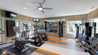 This fitness center offers a thoughtfully designed workout environment that combines functionality with an energizing aesthetic. The space features warm wood-paneled walls that lend a welcoming, casual feel, complemented by a striking multi-colored striped floor in shades of orange, gray, and teal that adds a dynamic visual appeal. A large ceiling fan keeps the air circulating throughout the room, while recessed lighting ensures the space is well-lit at all times.

The cardio section is well-appointed with multiple treadmills, elliptical trainers, and other aerobic machines, all arranged to allow comfortable use and easy navigation. A full-length wall mirror visually expands the space and allows users to monitor their form during workouts. Mounted flat-screen televisions are positioned for viewing from the cardio equipment, enhancing the exercise experience. A cable strength training machine rounds out the equipment offerings, providing options for resistance and strength workouts.

Large windows and glass-panel doors invite natural light into the space and offer views of the surrounding greenery outdoors, creating an open and refreshing atmosphere. Overall, this fitness center provides a clean, bright, and well-organized environment suited for a variety of workout routines.