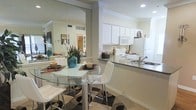 This single-floor residence offers a thoughtfully designed layout that seamlessly connects the kitchen and dining areas, creating an open and functional living environment. The kitchen is outfitted with white cabinetry that extends to the upper walls, providing generous storage space. A white appliance suite includes a smooth-top range, microwave, and refrigerator, all complementing the clean, classic aesthetic. Dark granite-style countertops add a refined contrast, while a dual-basin sink with a modern faucet sits along the breakfast bar peninsula, which also serves as a natural divider between the kitchen and dining space. The dining area comfortably accommodates a glass-top table with chrome accents and cushioned white chairs, centered on a patterned area rug that adds visual interest to the tiled flooring. A large wall mirror in the dining area amplifies the sense of space and reflects natural light throughout the room, enhancing the bright and airy atmosphere. Crown molding details along the ceiling add an elegant architectural touch. The overall ambiance is clean, modern, and inviting, making it an ideal space for both everyday living and casual entertaining.