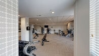 Located at 205 W Touhy Ave in Park Ridge, IL, this fitness facility spans a generously sized single floor divided into two dedicated exercise rooms, offering a thorough and well-organized workout environment. The space features a modern, clean aesthetic with neutral gray walls, drop-panel ceilings with recessed lighting, and a combination of durable carpet tile and smooth hardwood-style flooring throughout.

The strength training room is equipped with a power rack loaded with weight plates, a multi-functional cable machine, adjustable benches, and a variety of resistance machines targeting multiple muscle groups. Full-length mirrors line the walls, enhancing the sense of openness and allowing users to monitor their form. A built-in counter with cabinetry and a chalkboard-style display panel provide a functional station for tracking workouts or storing essentials.

The cardio room is equally well-appointed, featuring treadmills, elliptical trainers, a rowing machine, and stationary bikes, including a recumbent model. Wall-mounted flat-screen TVs offer entertainment during workouts, while natural light filters in through windows, creating a bright and motivating atmosphere. Large mirrors further amplify the room's spaciousness. Together, the two rooms deliver a comprehensive, amenity-rich fitness experience suited for a wide range of training goals.