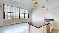 Located at 1 Upper Falls Rd. in Biddeford, Maine, this single-floor unit blends industrial character with modern finishes to create a clean, functional living space. Polished concrete floors run throughout, lending a raw, loft-like aesthetic that is complemented by soaring ceilings with exposed structural elements and track lighting.

The open-concept living and kitchen area serves as the heart of the unit. The kitchen is well-appointed with white shaker-style cabinetry, stainless steel appliances including a refrigerator and range, a tile backsplash, and a peninsula featuring dark stone countertops and a built-in sink — ideal for both cooking and casual dining. Stylish industrial pendant lights hang above, adding warmth to the space. Large, multi-pane windows flood the living area with abundant natural light and offer views of the surrounding greenery.

The bedroom is a comfortable, private retreat with neutral gray walls, clean white trim, and continued concrete flooring. It is accompanied by a dedicated closet for storage. A hallway connects the living spaces to the bedroom, maintaining a logical and efficient layout throughout.

The overall ambiance is modern and minimalist, with the industrial design elements giving the space a distinctive character well-suited to Biddeford's revitalized mill district setting.
