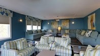 Located at 681 Darlington Avenue in Wilmington, North Carolina, this single-floor space offers a thoughtfully designed interior that blends comfort with contemporary style. The lounge area serves as the centerpiece of the space, showcasing rich teal-blue walls that establish a calming, coastal-inspired atmosphere. Large windows allow natural light to filter in, brightening the interior and complementing the cool color palette. The room is furnished with an array of plush seating options, including sectional sofas and accent chairs upholstered in coordinating teal and green-striped fabric, providing ample room for gathering and relaxation. Decorative wall art with soft, abstract botanical motifs adds an artistic focal point, while wall-mounted sconces contribute warm, ambient lighting throughout. Low-profile coffee tables are thoughtfully positioned throughout the seating arrangement to enhance functionality. The dark hardwood-style flooring provides a grounding contrast to the lighter furnishings and wall tones. Decorative valances over the windows add a refined finishing touch, tying together the cohesive interior design. Overall, the space exudes a welcoming and polished ambiance well-suited for communal use.