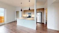 This single-floor unit showcases a clean, contemporary design with a consistent warm hardwood-style flooring and crisp white walls throughout. The open-concept kitchen serves as a central highlight, featuring light wood cabinetry extending to the ceiling, a white quartz countertop island with an undermount sink, stainless steel appliances including a range, microwave, and refrigerator, and a subway tile backsplash. Two sleek black pendant lights hang above the island, adding a modern touch. The kitchen opens into the surrounding living areas, creating a connected and functional layout.

The bedrooms are well-proportioned, each benefiting from large windows that invite abundant natural light and offer views of the surrounding urban environment. The rooms maintain a minimalist aesthetic with neutral tones and clean lines.

Two bathrooms serve the unit. One features a glass-enclosed walk-in shower, a single-sink vanity with light wood cabinetry, and chrome fixtures. The second bathroom includes a matching vanity and toilet, finished with the same consistent flooring. An in-unit laundry area is also visible within the layout. Overall, the space presents a bright, well-organized, and modern living environment.