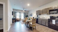 This single-floor residence offers a thoughtfully designed open-concept layout that seamlessly connects the kitchen, dining, and living areas into one cohesive and functional space. The interior is finished with warm wood-look luxury vinyl plank flooring throughout the main living areas, complemented by neutral wall tones that create a bright and welcoming atmosphere.

The kitchen is well-equipped with dark taupe cabinetry, black appliances including a refrigerator, electric range, microwave, and dishwasher, along with a double-basin sink. The dining area sits adjacent to the kitchen, offering convenient access for everyday meals. The living room is generously sized, featuring a ceiling fan, a wall-mounted television, and direct access to an outdoor patio through glass doors framed by bold blue-and-white patterned curtains that add a stylish coastal flair. Natural light flows freely through the large windows and patio doors.

The bedroom is a serene retreat with plush carpeting, a large window with blinds and sheer curtains, and vaulted ceiling details that add architectural interest. The full bathroom includes a dark-toned vanity with ample cabinetry, a large mirror, a shower-tub combination, and wood-look flooring. An open shelving area adjacent to the bathroom provides additional storage.