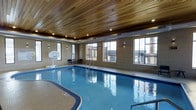 This single-floor indoor recreation facility is centered around a generously sized swimming pool with clear blue water, complete with stainless steel entry railings for safe access. The pool is framed by tile-finished walls and a striking warm wood-paneled ceiling fitted with recessed pot lights, creating an inviting and upscale ambiance. Wall-mounted sconce lighting complements the natural light that pours in through a series of large, multi-pane windows lining the perimeter, offering views of the surrounding exterior. A basketball hoop is mounted along one wall, adding a recreational dimension beyond aquatic use. Seating is available along the poolside, providing a comfortable spot for spectators or those taking a break. Exposed ductwork along the upper walls adds an industrial accent that contrasts tastefully with the warmth of the wood ceiling. The overall design balances functionality and aesthetics, making it well-suited for both fitness and leisure activities. The space is clean, modern, and thoughtfully appointed, reflecting a high standard of recreational amenity design.
