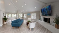 Located at 1699 E Washington St in Colton, California, this single-story space delivers an open, airy atmosphere defined by its striking vaulted ceilings with exposed beams and recessed lighting throughout. Light-toned luxury vinyl plank flooring runs seamlessly across the entire space, enhancing the sense of continuity and openness.

The expansive main living area is a standout feature, anchored by a white shiplap fireplace wall with a large wall-mounted display above it. A tufted blue sofa, paired with white accent chairs and a wooden coffee table, creates a comfortable and stylish seating arrangement. Generous windows with horizontal blinds line multiple walls, filling the space with natural light and offering views of the surrounding greenery.

A dedicated dining area sits adjacent to the living space, benefiting from its own bank of windows and access to the exterior through glass French doors, allowing for excellent indoor-outdoor connectivity.

The kitchen is well-appointed with crisp white cabinetry, white subway tile backsplash, stainless steel appliances including a refrigerator, range, and microwave, as well as a stainless undermount sink. The clean, neutral palette throughout the kitchen complements the overall modern aesthetic of the space.

Together, these areas form a cohesive, light-filled environment that is both functional and welcoming.