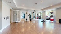 Located at 1055 Hatches Pond Lane in Cary, NC, this single-floor fitness facility offers a thoughtfully designed space for comprehensive health and wellness activities. The layout is divided into distinct functional zones, including a spacious exercise room and a living room area, all finished with warm hardwood-style flooring and a clean, neutral color palette throughout.

The exercise room is the centerpiece of the facility, featuring floor-to-ceiling windows that flood the space with natural light and provide views of the surrounding greenery. Full-wall mirrors amplify the sense of openness. The room is outfitted with a wide array of equipment, including treadmills, an elliptical, a stationary bike, a rowing machine, a cable resistance machine, and an assortment of free weights — from dumbbells to medicine balls and exercise balls. Dedicated storage racks keep equipment organized and accessible.

A separate multi-purpose area features light hardwood flooring, recessed lighting, a wall-mounted display screen, and built-in cabinetry with a beverage station — ideal for stretching, yoga, or guided workout sessions. Glass-panel doors connect the zones, maintaining an open, airy flow throughout. Tray ceilings with recessed lighting add architectural refinement, making this facility as visually polished as it is functionally complete.