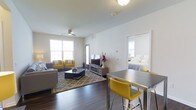 Located at 199 Cedar Park Boulevard in Northampton County, Pennsylvania, this single-floor residence offers a well-designed, open-concept layout that seamlessly connects the main living spaces. The living room is bright and welcoming, featuring rich dark hardwood flooring, large windows that invite generous natural light, and a ceiling fan overhead. The space is thoughtfully arranged with comfortable seating, a media console with a flat-screen television, and a patterned area rug that anchors the room. Adjacent to the living area, a dining space with a raised bar-height table and bold yellow bar stools provides a functional and stylish spot for meals. The open sightlines between these areas create a sense of spaciousness throughout. A glimpse into the bedroom reveals a neatly appointed space with neutral tones, natural light streaming through a window, and coordinating accents that tie into the home's cohesive color palette of grays, whites, and yellow. The clean white walls and smooth ceilings throughout contribute to a fresh, modern aesthetic. Overall, the residence presents a comfortable and practical living environment with contemporary finishes and a bright, airy ambiance.