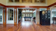 This expansive single-floor clubhouse is a masterfully designed community gathering space that blends rustic charm with refined comfort. Dramatic vaulted ceilings with exposed dark wood timber trusses define the architectural character, soaring overhead and creating an airy, grand atmosphere throughout the main areas. Rich hardwood flooring runs continuously across the space, anchoring each zone with warmth and cohesion.

The main lounge area is anchored by a striking floor-to-ceiling stacked stone fireplace, flanked by built-in shelving and comfortable wingback seating — an ideal spot for relaxed conversation. Adjacent, the game room impresses with a full-size billiards table and foosball table, bathed in natural light pouring through large multi-pane French doors and clerestory windows that overlook lush greenery.

A welcoming lobby features an open seating arrangement and views that draw the eye toward the outdoors through walls of glazed French doors. The business center offers a dedicated workstation area alongside casual lounge seating, with direct access to an outdoor terrace. A transitional corridor connects to a fitness area, finished with decorative area rugs and full-length mirrors. Throughout, generous window placement ensures each area is filled with natural light, while the cohesive use of wood tones and neutral palettes creates a polished, inviting ambiance.