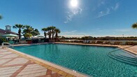 Located on Floral Springs Boulevard in Port Orange, Florida, this outdoor amenity complex offers a well-appointed resort-style pool environment designed for relaxation and recreation. The centerpiece is a generously sized, rectangular swimming pool with crystal-clear turquoise water, surrounded by a broad, multi-toned paver deck that provides ample space for lounging. Numerous poolside chaise lounges are arranged along the perimeter, offering plenty of seating options under the abundant Florida sunshine.

A separate spa or wading pool is positioned near the main structure, accompanied by shaded seating areas with patio tables and umbrellas in a distinctive teal and white striped pattern. A charming gazebo with a hip roof serves as an additional shaded gathering spot within the fenced enclosure.

The adjacent clubhouse building is designed in a classic Florida vernacular style, featuring a hipped roof with a cupola, white columns, large windows, and a covered veranda. Lush tropical landscaping—including tall palms, flowering shrubs, and colorful plantings—frames the entire area, creating a vibrant, resort-like atmosphere. The pool deck offers open views toward a waterway beyond the perimeter fence, enhancing the overall serene, outdoor ambiance of this community amenity space.