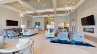 Located at 2600 Executive Dr in Venice, Florida, this upscale amenity clubhouse delivers a refined, resort-inspired experience through thoughtful architectural design and high-end finishes. The grand entrance corridor sets the tone immediately, featuring large-format tile flooring, a dramatic floor-to-ceiling water wall feature, and a stylish reception desk with a dark stone countertop and natural wood paneling. Overhead, a sculptural gold chandelier and recessed coffered ceiling details add warmth and elegance.

The space opens into expansive lounge areas bathed in natural light from floor-to-ceiling windows and glass doors that frame views of the outdoor surroundings, including a pool area and lush Florida landscaping. Soaring coffered ceilings with modern sarburst chandeliers lend a sense of grandeur, while the neutral tile floors and soft color palette of creams, blues, and grays create a serene, contemporary atmosphere.

Comfortable seating arrangements with plush sofas and accent chairs are thoughtfully grouped throughout, complemented by a textured 3D accent wall with a sleek linear fireplace and a wall-mounted television. Sliding barn doors and glass-paned French doors provide access to additional areas. Secure keypad entry points ensure controlled access throughout the facility.