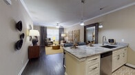 This thoughtfully designed single-floor residence offers an open-concept layout that seamlessly connects the kitchen, dining, and living areas. The kitchen is appointed with granite countertops, cream cabinetry, stainless steel appliances including a refrigerator and range, a subway tile backsplash, and an island with pendant lighting overhead — ideal for both cooking and casual dining. Dark hardwood-style flooring flows through the main living spaces, complementing the warm, neutral color palette throughout.

The living room features crown molding, a ceiling fan, and large windows dressed with elegant patterned drapes that invite natural light. A striking gold geometric decorative panel serves as a focal point, adding a contemporary flair to the space.

The primary bedroom is generously sized with plush carpet, a ceiling fan, ample natural light from multiple windows, and stylish furnishing potential. A second bedroom mirrors this comfort with similarly soft carpeting and bright windows. A versatile flex room provides additional space suitable for a home office or guest room.

Both bathrooms are finished with granite-topped vanities, cream cabinetry, framed mirrors, and dark hardwood-style flooring, offering a refined and cohesive aesthetic. Throughout the residence, crown molding, quality finishes, and a warm neutral palette create an inviting and polished atmosphere.