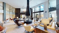 This striking lobby space exemplifies contemporary design through a carefully considered blend of architectural drama and refined furnishings. The centerpiece of the main lounge is a striking suspended conical fireplace finished in matte black, positioned above a circular base and surrounded by an eclectic arrangement of seating that includes sculptural armchairs, plush sofas, and woven accent chairs atop a vivid abstract area rug. Large floor-to-ceiling windows flood the space with natural light while framing outdoor views.

Dark wood wall paneling, accented by a geometric brass lattice feature wall, adds warmth and visual texture throughout. A sweeping matte black spiral staircase serves as a bold sculptural element, connecting areas and adding vertical drama to the double-height volume. The reception desk area features fluid, organic forms in dark metal with brass accents.

Secondary seating alcoves offer more intimate settings, furnished with leather banquettes and accent chairs in warm tones. Pale large-format stone tile flooring flows throughout, grounding the space and complementing the neutral-toned walls. Recessed lighting and curated table lamps contribute to a layered, inviting ambiance suited to both social gathering and quiet relaxation.