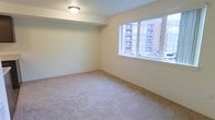 This single-floor residential space offers a clean and straightforward layout designed for comfortable everyday living. The main room is finished with wall-to-wall neutral-toned carpeting that adds warmth and softness underfoot, while smooth, light-colored walls create a bright and airy atmosphere throughout. A large window is a standout feature of the space, flooding the interior with abundant natural light and framing views of the neighboring urban buildings and streetscape beyond. The window's generous proportions enhance the sense of openness, making the room feel welcoming and connected to its surroundings. Ceiling height is standard, with a flush-mounted ceiling light fixture providing practical overhead illumination. The walls are kept clean and simple, offering a neutral canvas well-suited to a variety of décor styles. The overall ambiance of the space is calm and functional, with an emphasis on light and simplicity. Its straightforward design makes it highly adaptable, whether used as a living area, bedroom, or flexible multi-purpose room. The urban views visible through the window situate this space within a lively neighborhood setting.