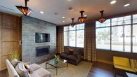This thoughtfully designed single-floor space blends contemporary style with warm, inviting elements to create a comfortable and functional lounge environment. The main seating area is anchored by a striking floor-to-ceiling tile accent wall that frames a sleek linear fireplace and a wall-mounted flat-screen television, making it an ideal focal point for gathering and relaxation. Rich hardwood flooring runs throughout, complementing the warm, neutral tones of the walls and furnishings.

A series of distinctive Craftsman-inspired pendant light fixtures with amber stained-glass shades hang from the ceiling, adding character and a warm glow to the space. Recessed lighting provides additional illumination, ensuring the area is well-lit at all times. An expansive bank of large windows along one wall floods the interior with generous natural light and offers views of the surrounding streetscape, dressed with striped drape panels for a polished finish.

Seating is arranged thoughtfully, featuring a plush dark sofa, accent chairs in bold yellow, and a glass-top coffee table with a metallic frame. An adjacent kitchenette area provides wood cabinetry, dark countertops, stainless steel appliances, and a sink — offering practical amenities for entertaining or daily use. The overall ambiance is sophisticated yet approachable, balancing modern design with welcoming comfort.