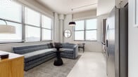 This single-floor unit presents a cohesive modern aesthetic defined by a neutral palette of whites and grays, light concrete-style flooring, and an abundance of natural light streaming through large, horizontally slatted windows throughout. The living area serves as the centerpiece of the space, furnished with a generously sized sectional sofa in steel-blue velvet, anchored by a textured area rug and a sculptural pedestal side table. A pendant light fixture hangs overhead from a wood-paneled ceiling, adding warmth to the otherwise crisp interior. A compact dining area with a small table and chair sits adjacent, making efficient use of the open-plan layout. A wall-mounted flat-screen television and media console complete the living zone. The kitchen is outfitted with floor-to-ceiling cabinetry in a warm gray wood-grain finish, stainless steel appliances including a full-size range and side-by-side refrigerator, and under-cabinet lighting that highlights the clean countertops. The bedroom accommodates a full-size bed framed by large windows offering urban views, with a padded bench at the foot providing a finishing touch. The bathroom features mosaic tile flooring, a combination tub and shower with full tile surround, and a streamlined vanity.