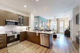Located at 12500 Live Oak Way in Frisco, TX, this single-floor residence offers 2 bedrooms, 2 bathrooms, and a thoughtfully designed open-concept layout. The combined kitchen and living area serves as the heart of the home, anchored by a sleek kitchen featuring rich dark wood cabinetry, a full suite of stainless steel appliances — including a French door refrigerator, range, microwave, and dishwasher — and a granite-topped island with bar seating. A white subway tile backsplash adds a clean, modern contrast. Light wood-style flooring flows throughout the main living area, which is brightened by large windows and a glass door opening to a private balcony. The living room is highlighted by a soft teal accent wall and a ceiling fan, creating a relaxed yet stylish ambiance. The primary bedroom features plush carpet, a bold accent wall, a ceiling fan, and generous natural light from a large window with outdoor views. The second bedroom is similarly carpeted and well-lit. Both bathrooms are finished with granite countertops, dark wood vanities, and built-in shelving. Additional spaces include two closets, a dedicated laundry area, and a utility room for added functionality.