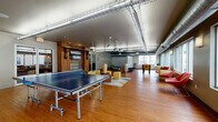 Located at 80 E Exchange St in downtown Akron, Ohio, this single-floor recreational lounge — aptly identified as "The Depot" — blends industrial design with a lively, social atmosphere. The open-plan layout is anchored by warm hardwood flooring that runs throughout the generous space, complemented by exposed ductwork and a mix of pendant and fluorescent lighting that give the area an urban, loft-like character.

A standout architectural feature is a preserved wood-paneled fireplace surround with brick detailing and a built-in bench, serving as a rustic focal point that adds historical charm to the otherwise modern setting. A wall-mounted flat-screen television is integrated into this structure, making it a natural gathering hub.

The recreational zone houses multiple ping-pong tables and a foosball table, offering ample space for active play. A lounge area along the windowed perimeter features bold red accent chairs and patterned ottomans arranged to take advantage of the natural light streaming through large windows that overlook the surrounding cityscape. Additional seating and activity stations are distributed throughout, including a giant Jenga set and other social games.

Access to the floor is provided via a metal-railed staircase near the "DEPOT" signage. The overall ambiance is energetic yet comfortable, making this space well-suited for community recreation and social engagement.