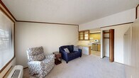 Located at 103 Jasmine Dr in Rocky Mount, NC, this efficient single-floor space is thoughtfully laid out to maximize functionality across a living room, kitchen, and bathroom. The living room is carpeted and features a textured ceiling with crown molding, a large front-facing window that brings in natural light and offers views of the surrounding greenery, and a built-in wardrobe unit with ample storage drawers along one wall. A front entry door with a sidelight panel provides easy access and additional natural light. Adjoining the living area is a built-in desk and open shelving unit, creating a seamless transition into the kitchen zone. The kitchen is equipped with light wood cabinetry, a white electric range, a stainless steel dishwasher, and tile flooring. A built-in countertop peninsula provides additional workspace and storage. The bathroom features tile flooring, a wood vanity with a granite-style countertop, and a clean, functional layout. Throughout the space, warm wood tones in the cabinetry and trim work complement the neutral wall colors, creating a cohesive and practical interior well-suited for everyday living.