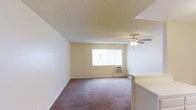 This single-floor space offers a straightforward and functional layout designed for comfortable everyday living. The main living area is finished with wall-to-wall carpeting in a neutral tone, providing a warm and quiet underfoot experience. Soft, cream-colored walls contribute to a clean and open ambiance throughout the room. A ceiling fan with an integrated light fixture is centrally positioned, offering both air circulation and overhead lighting. Natural light enters the space through a window dressed with vertical blinds, which also houses a wall-mounted air conditioning unit for climate control. A textured popcorn ceiling adds a classic finishing detail overhead. A partial countertop or partition is visible along one edge of the room, suggesting an adjacent kitchen or bar area that provides a subtle sense of division between living and utility spaces. The overall layout is open and unobstructed, making the space highly adaptable to a variety of furniture arrangements and uses. The neutral color palette and practical finishes give the space a versatile, move-in-ready character suited to a range of occupants.