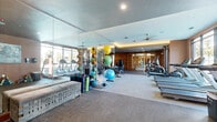 This modern fitness center is thoughtfully designed across a single floor, offering two distinct zones that cater to a wide range of workout preferences. The main exercise area features a lineup of Matrix-brand cardio machines — including treadmills, ellipticals, and a stair climber — positioned along floor-to-ceiling windows that flood the space with natural light and provide views of the surrounding outdoors. A row of strength training machines lines a striking warm wood-paneled accent wall, creating a polished, contemporary aesthetic. Floor-to-ceiling mirrors amplify the sense of space and allow exercisers to monitor their form throughout their workout. A functional training zone is stocked with medicine balls, stability balls, foam rollers, resistance equipment, and adjustable benches, supported by additional mirror panels. A padded bench and a built-in beverage cooler offer added convenience. The second zone serves as a dedicated studio or group fitness room, featuring a bold hexagonal-patterned carpet, warm wood walls, ceiling fans, and large windows with outdoor views. Wall-mounted speakers, mirrors, and a collection of stability and medicine balls make it ideal for classes or independent training sessions.
