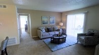 This single-floor residence offers a thoughtfully arranged living space designed for both comfort and functionality. The living room serves as the heart of the home, featuring plush wall-to-wall carpeting in a neutral tone that adds warmth underfoot. Soft, beige-toned walls create a calming backdrop throughout the space, complemented by a textured ceiling that adds subtle architectural character. A large sliding glass door with vertical blinds floods the room with natural light and provides direct access to an outdoor area, enhancing the sense of openness. The living area is well-proportioned, offering ample room for comfortable seating arrangements centered around a oval glass-top coffee table. A doorway visible from the living room connects to additional areas of the residence, suggesting a practical and flowing floor plan. Decorative wall art featuring coastal-inspired imagery adds a serene, relaxed aesthetic to the space. Overall, the residence presents a clean, move-in ready environment with a cohesive design language — neutral tones, soft textures, and generous natural light — making it an inviting and livable space on a single, accessible level.