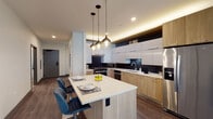 Located in Chicago's vibrant Loop neighborhood, this well-appointed single-floor residence showcases a thoughtfully designed modern interior. The open-concept kitchen is a standout feature, boasting a two-tone cabinetry scheme that pairs warm wood-grain lower cabinets with sleek white upper cabinets, all set against a striking dark herringbone tile backsplash. A generous island with a white countertop and blue upholstered bar seating serves as both a dining and gathering space, illuminated by a trio of stylish pendant lights. Stainless steel appliances, including a French door refrigerator, gas range, dishwasher, and microwave, complete the well-equipped culinary space. The living area transitions seamlessly from the kitchen, featuring wide-plank hardwood flooring, large floor-to-ceiling windows that frame city views, and ample natural light. A dedicated reading nook with a window overlooking the surrounding Chicago architecture adds a charming retreat within the space. The bedroom offers a calm, carpeted environment with soft gray walls, a large window, and warm wood furniture. The bathroom is finished with large-format tile flooring, a floating wood vanity, and clean, minimalist fixtures, reflecting the contemporary design language found throughout the residence.