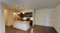 Located in Midland, this single-floor space offers a thoughtfully designed open-concept layout that seamlessly connects the kitchen and living areas. The kitchen is a standout feature, showcasing dark espresso cabinetry that contrasts elegantly with light granite countertops. A central island with a built-in sink provides additional workspace and serves as a natural gathering point. Stainless steel appliances, including a refrigerator, range, and microwave, complete the modern culinary setup. Pendant lighting above the island adds warmth and definition to the space, while track lighting throughout ensures flexible and ample illumination. Rich, dark hardwood flooring runs throughout, grounding the space with a sense of warmth and continuity. The open living area beyond the kitchen offers generous room to arrange furnishings and personalize the space to suit any lifestyle. Crown molding along the ceiling adds a refined architectural touch, elevating the overall aesthetic. The layout balances both functionality and style, making it an inviting and practical space for everyday living.