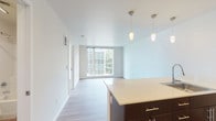 This well-appointed single-floor residence showcases a clean, contemporary design throughout. The open-concept layout seamlessly connects the kitchen and living area, creating a bright and functional shared space. Large floor-to-ceiling windows flood the living room with natural light and frame views of the surrounding urban landscape, while a sliding glass door provides additional access to the outdoors.

The kitchen is a standout feature, centered around a substantial island with a light-toned countertop, undermount sink, and sleek dark cabinetry with brushed silver hardware. Pendant lighting hangs above the island, complementing the recessed lighting above. White appliances, including a refrigerator, stove, and microwave, are neatly arranged along the galley-style wall cabinetry.

Light-toned wood-look flooring runs consistently throughout the unit, lending warmth and cohesion to every room. The bedrooms are generously proportioned, with clean white walls and ample natural light, one of which benefits from a large window wall overlooking the city.

Both bathrooms follow the same design language, featuring dark wood vanities with white countertops, single-basin sinks, large mirrors, and a tub-shower combination. The overall palette of white walls, light floors, and dark cabinetry accents creates a modern, fresh, and inviting atmosphere throughout.