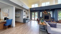 This well-appointed community clubhouse or leasing lounge presents a thoughtfully designed single-floor layout that balances functional and social spaces with a cohesive, modern aesthetic. The interior is defined by striking slate-blue accent walls contrasted against crisp white upper walls and trim, creating a bold yet inviting atmosphere throughout.

Soaring ceilings with clerestory windows allow natural light to pour in from multiple angles, enhancing the sense of openness and airiness in the main gathering area. Warm-toned wood-look flooring runs continuously throughout the space, grounding the design with a welcoming feel.

The main lounge area features a stone fireplace flanked by decorative wall accents and comfortable seating, making it an ideal space for relaxed gatherings. A built-in kitchenette with teal cabinetry, open shelving, and subway tile backsplash provides a convenient refreshment station. Large glass double doors open to an outdoor terrace or pool area, blurring the boundary between interior and exterior.

The entryway is welcoming and functional, with a glass-paneled entry door, entry mat, and a tufted bench seating area. Throughout the space, curated artwork — including a large abstract canvas in warm gold tones — adds a polished, gallery-like quality. Bistro-height seating, accent chairs in rich cognac leather and deep blue velvet, and wooden tables complete the stylish yet comfortable furnishings.