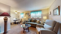 This well-appointed single-floor residence offers a cohesive and comfortable layout with a warm, coastal-inspired color palette running throughout. The open living and dining area serves as the heart of the home, featuring plush carpeted flooring, multiple windows dressed in vibrant teal curtains that invite natural light, and a ceiling fan overhead. The living space is thoughtfully arranged with seating areas, a glass-top coffee table, and a dedicated media corner, creating distinct yet connected zones for relaxing and entertaining. A dining area with seating for several occupants sits just off the living room, making the space ideal for everyday use. The kitchen is efficiently designed with warm oak cabinetry, a tile backsplash, tile flooring, and white appliances including a gas range and refrigerator, offering ample storage and counter space. The bedroom is a standout feature, characterized by a bold yellow accent wall that adds energy and warmth to the room, complemented by soft carpeting, a large sleigh bed, and a window with garden views. The bathroom continues the aquatic theme with blue tile work and coordinating accessories. Throughout the home, consistent décor choices in teal and yellow tones create a unified and inviting atmosphere.