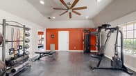 This fitness center is thoughtfully laid out across two interconnected rooms, offering a well-rounded environment for both cardio and strength training. The main workout room is immediately distinguished by its bold burnt-orange accent walls, which energize the space and set a motivating tone. Vaulted ceilings with a large ceiling fan and recessed lighting enhance the sense of openness and airflow, while durable polished concrete flooring throughout provides a practical, low-maintenance surface ideal for high-activity use. This room is equipped with a multi-functional cable machine, a weight bench, and a rack of dumbbells and stability balls with resistance bands, catering to a full range of strength and functional training exercises. A window along one wall admits natural light, adding brightness to the interior. The second room serves as a dedicated cardio zone, featuring multiple treadmills and elliptical machines arranged to face large windows with views of an outdoor pool area, creating an inviting atmosphere for sustained cardio sessions. A full-length mirror along one wall amplifies the sense of space and allows users to monitor their form. French doors in one area provide additional natural light and outdoor access. Together, the two rooms form a comprehensive, well-designed fitness facility suited for a diverse range of workout routines.